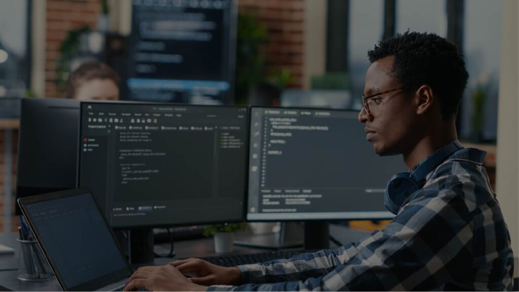 Développeur de logiciels tapant sur un ordinateur portable, assis à un bureau avec plusieurs écrans et en train d'analyser du code